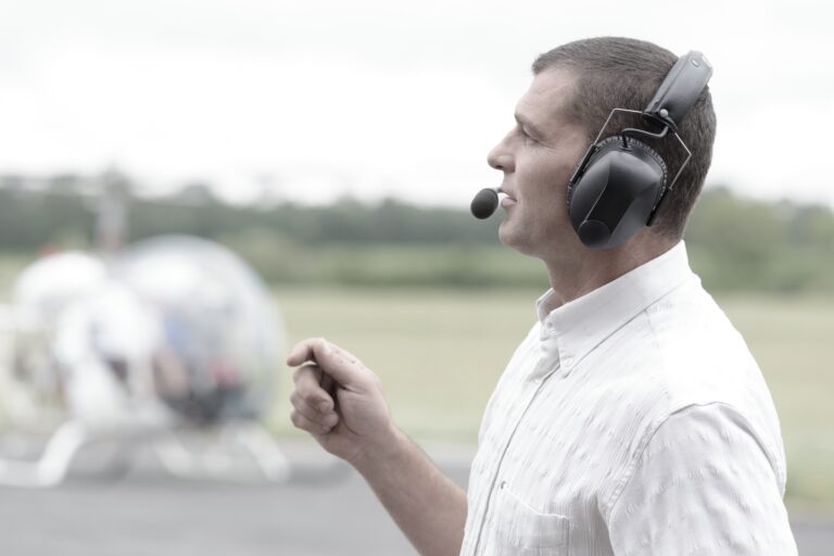 A man in a white shirt wearing a headset with a microphone stands outdoors, speaking or giving instructions. A helicopter is visible in the blurred background on a grassy area.