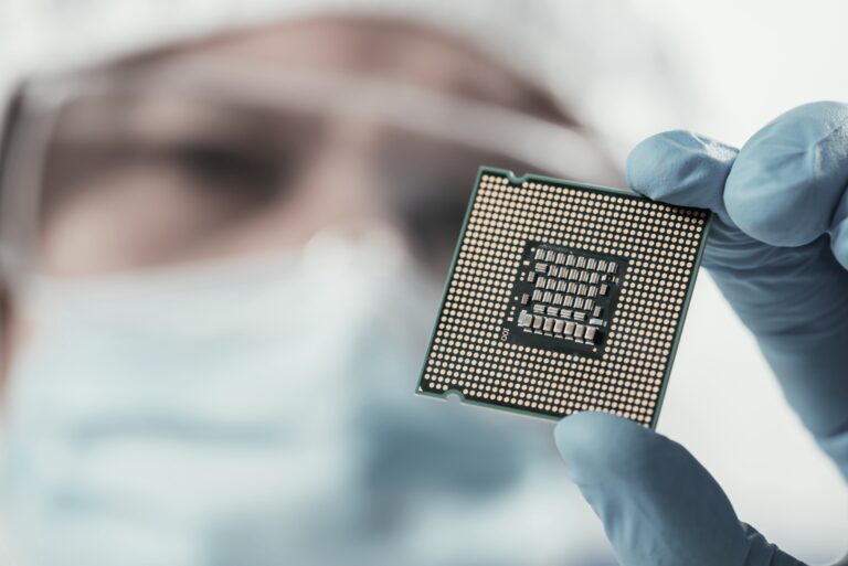 A person wearing a mask, gloves, and protective eyewear holds a computer processor chip between their fingers, with the focus on the chip and the person's face blurred in the background.