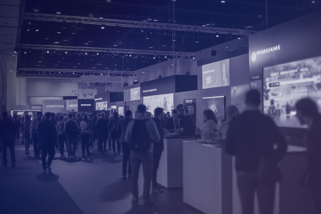 A group of people walks and stands among large display stands at an indoor exhibition or trade fair, with overhead lights and digital screens visible in the background.