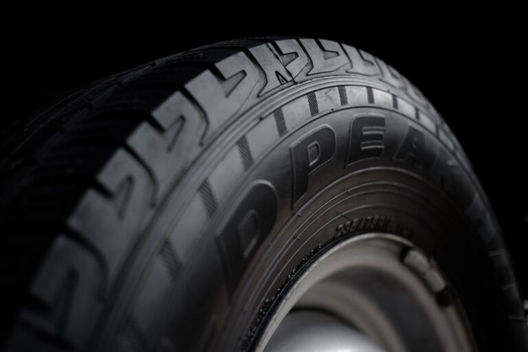 Close-up view of a black car tyre showing detailed tread pattern and partial sidewall text, mounted on a metal wheel rim, against a dark background.