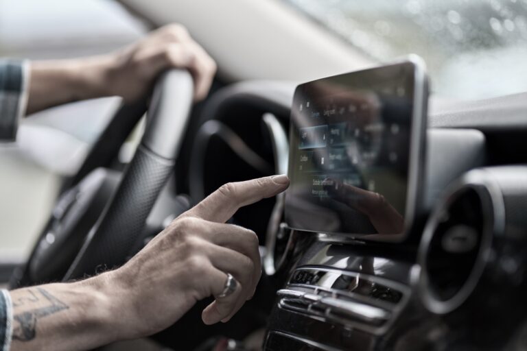 A person’s hand is touching the touchscreen of a car’s infotainment system while their other hand is on the steering wheel. The car interior and dashboard controls are visible.
