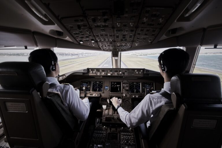 Two pilots in uniform sit in the cockpit of an aeroplane, both wearing headsets and facing the runway as they prepare to land the aircraft. The control panels and cockpit instruments are visible.