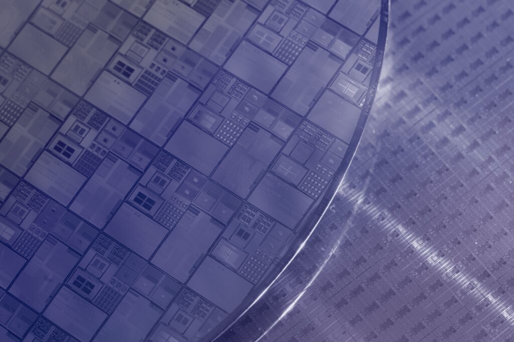 Close-up view of a silicon wafer showing multiple microchips with intricate geometric patterns, placed on a similarly textured surface. The image has a bluish-purple tint.