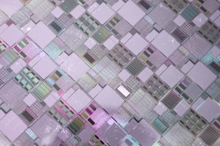 A close-up view of numerous rectangular and square microchips or integrated circuits, arranged in a grid pattern with reflective, metallic surfaces in shades of pink, green, and silver.