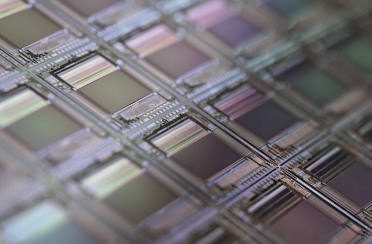 Close-up view of a silicon wafer with multiple microchips, showing intricate geometric patterns and reflective surfaces in muted shades of purple, green, and grey.