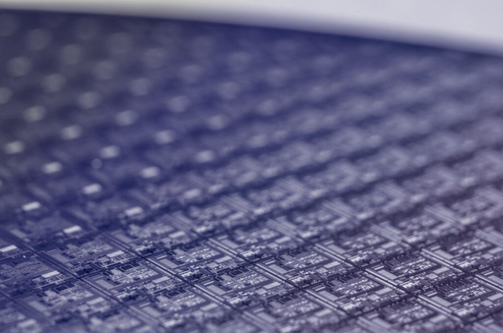 A close-up view of a silicon wafer covered with multiple integrated circuits, showing the detailed and repetitive pattern of electronic components. The image is in cool blue and purple tones.