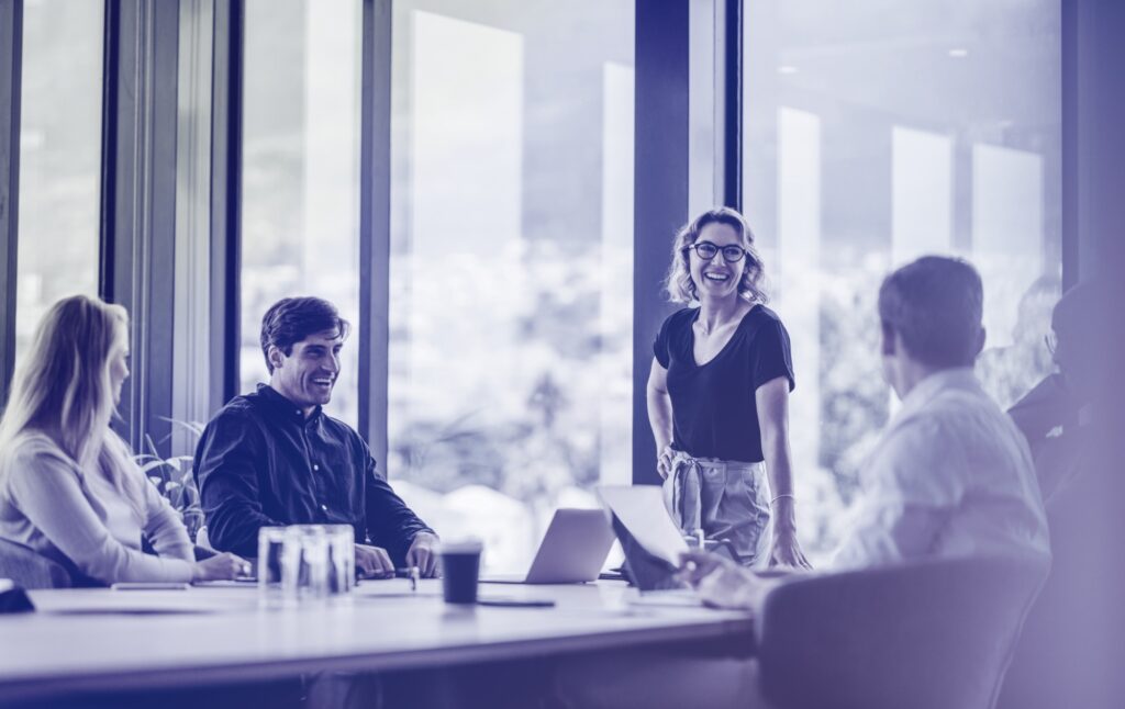 Four people are seated at a conference table while one person stands and speaks to the group. Large windows provide natural light, and laptops and glasses of water are on the table.