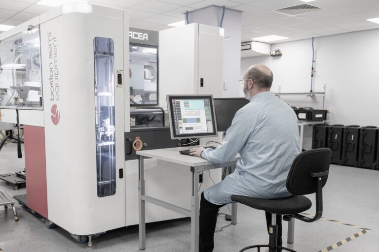 A man in a lab coat sits at a desk operating a computer in front of large laboratory equipment inside a clean, modern lab environment.