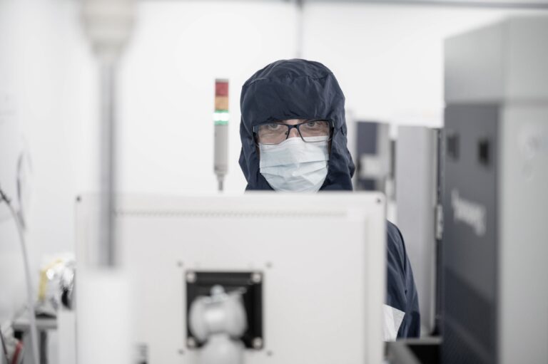 A person wearing a dark hooded overall, glasses, and a face mask looks at a computer monitor in a sterile, laboratory-like environment with technical equipment visible.