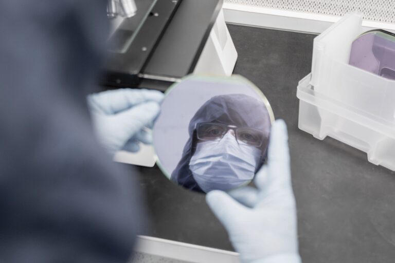 A person wearing a protective suit, mask, and gloves holds a reflective silicon wafer, showing their face reflected in it inside a laboratory setting.