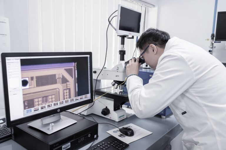 A person in a white lab coat looks into a microscope at a laboratory workstation, with a computer monitor displaying a magnified image of an electronic chip beside them. Various lab equipment surrounds the area.