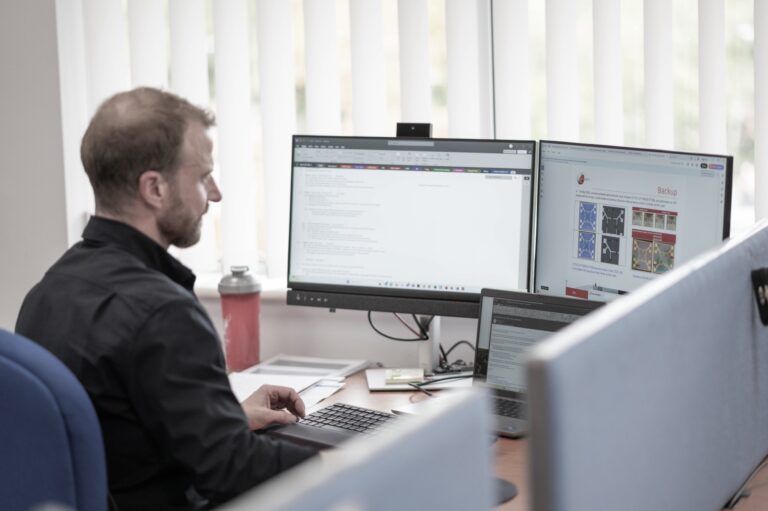 A man sits at a desk working on a computer with two monitors and a laptop. The screens display documents and a webpage. Office supplies and a water bottle are on the desk. White vertical blinds cover the window.