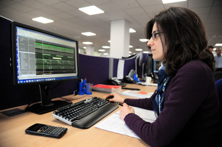 A person with long dark hair and glasses is working at a desk, looking at a computer monitor displaying audio editing software. A keyboard, calculator, notepad, and office supplies are on the desk.