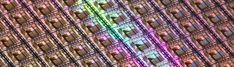 A close-up view of a silicon wafer showing multiple rectangular microchips with reflective surfaces, illuminated by green and pink light.